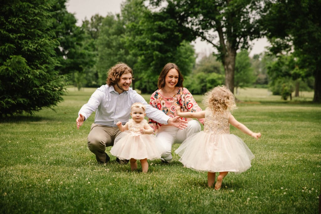 Candid photo of family of 4 laughing and smiling naturally with the parents arms outstretched and the little children running to give them a hug in a Cedar Rapids park.