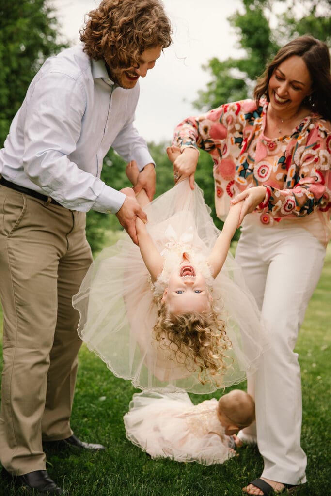 Fun, candid, photo of parents swinging their young daughter upside down while she giggles during a Spring lifestyle family photo session in an Eastern Iowa park.