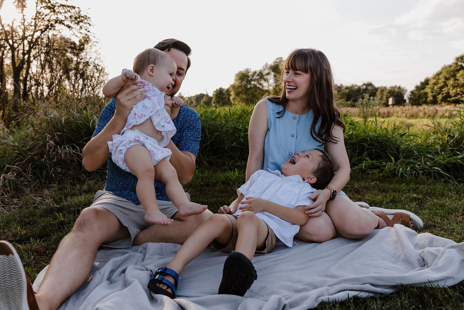 Family of 4 sitting on blanket laughing together during a candid family session in Cedar Rapids, Iowa park.