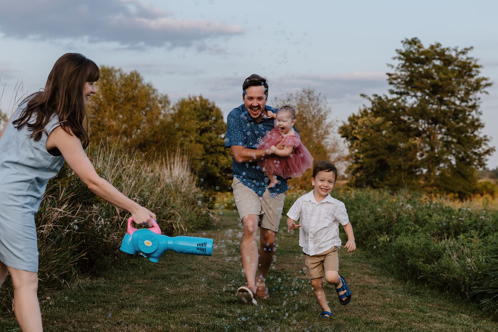 Candid moment of a family of 4 laughing together as mother holds bubble machine and father and two kids run through the bubbles laughing during a lifestyle family photo session in a park near Cedar Rapids, Iowa.