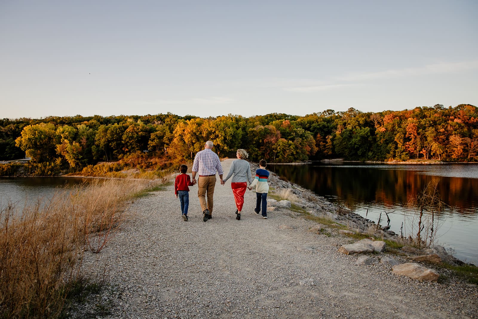 Family of 4 holding hands walking holding hands with bright fall foliage in the background during sunset the lake in North Liberty, Iowa.